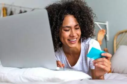 Happy young African American woman online shopping using laptop and credit card on bed in bedroom.