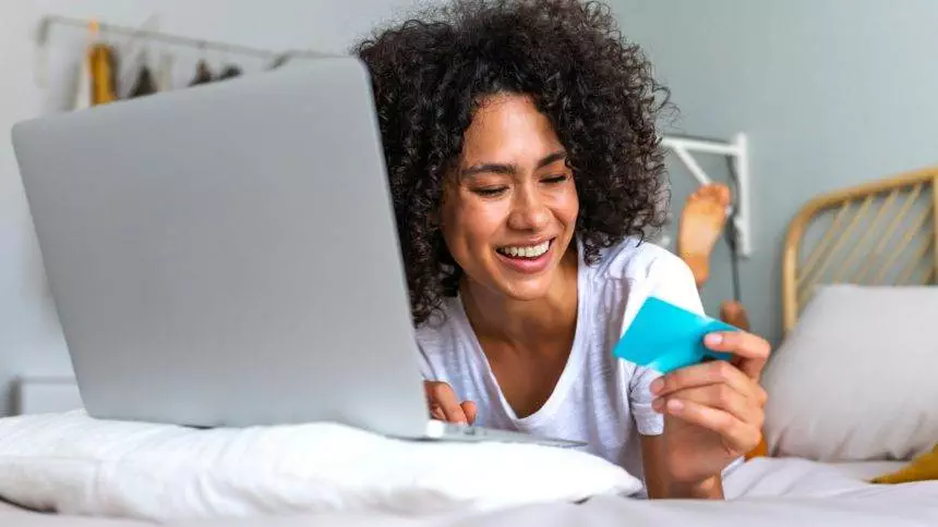 Happy young African American woman online shopping using laptop and credit card on bed in bedroom.